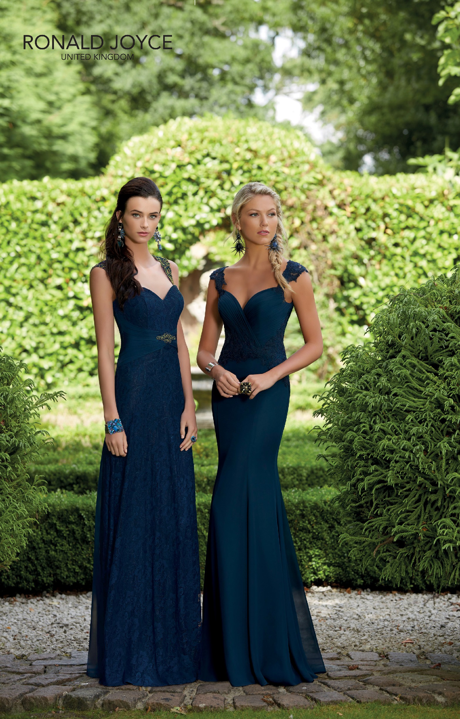 Two teenage bridesmaids stood in a posh garden. One has long dark brown hair and one has long blonde hair. Both wear dark green long bridesmaid dresses with sweetheart necklines